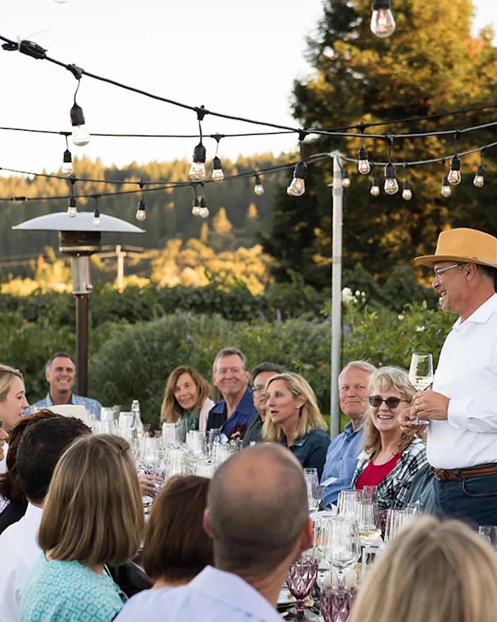 A leader addressing a long outdoor table of executives under string lights at dusk.